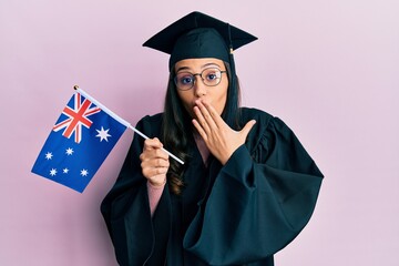 Young hispanic woman wearing graduation uniform holding australia flag covering mouth with hand, shocked and afraid for mistake. surprised expression