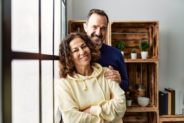 Middle age hispanic couple smiling happy leaning on the window at home.