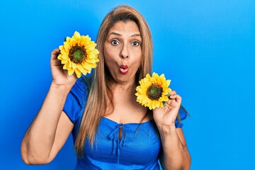 Middle age hispanic woman holding yellow sunflowers afraid and shocked with surprise and amazed expression, fear and excited face.