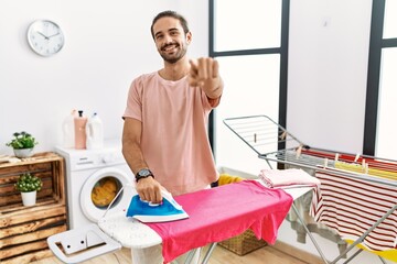 Obraz premium Young hispanic man ironing clothes at home pointing to you and the camera with fingers, smiling positive and cheerful