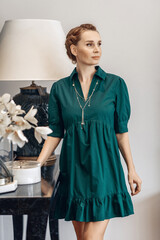 Portrait of adorable cute well-groomed woman wearing green dress and jewelry with neat hairdo posing next to a table and posing. Comfort concept