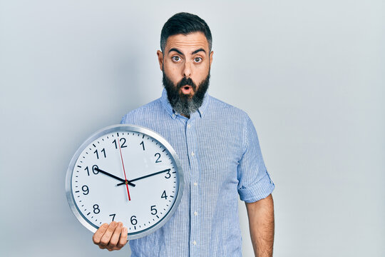 Young hispanic man holding big clock scared and amazed with open mouth for surprise, disbelief face