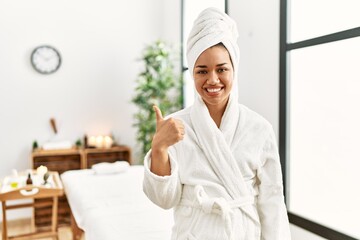 Young brunette woman wearing towel and bathrobe standing at beauty center doing happy thumbs up gesture with hand. approving expression looking at the camera showing success.