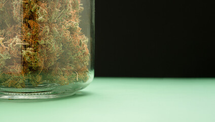 Dry cannabis buds flower in a jar glass on a green floor with a black background.