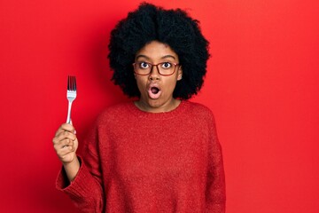 Young african american woman holding one silver fork scared and amazed with open mouth for...