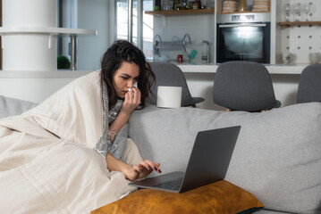 Young ill business woman wrapped in a blanket works on a laptop from home. Female college student sick with flu with common cold having online education class via video call on her computer.