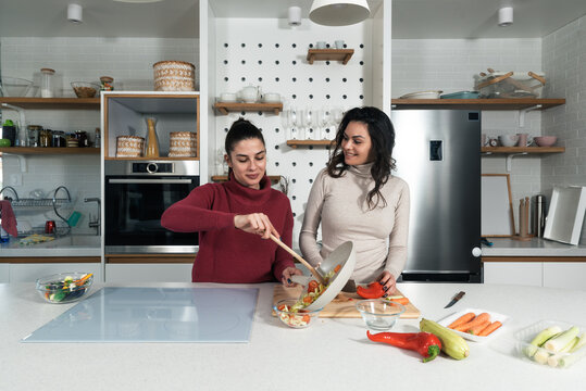 Two Young Happy College Student Roommates Or Business Women Cooking Food Together At Their Apartment. Females Having Fun Together Preparing Meal In Their Modern Kitchen At Cozy Home.