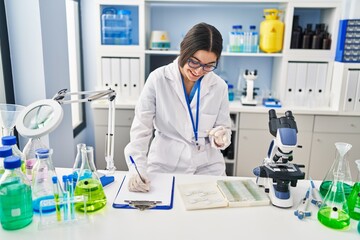 Young hispanic woman wearing scientist uniform writing on clipboard holding sample at laboratory