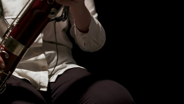 Woman sitting on chair in studio with bassoon on her lap picks it up and starts playing music, front view. Female musician plays bassoon at dark background