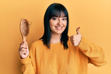 Young hispanic woman holding comb loosing hair smiling happy and positive, thumb up doing excellent and approval sign