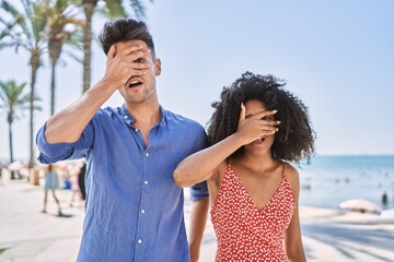 Young interracial couple outdoors on a sunny day peeking in shock covering face and eyes with hand, looking through fingers with embarrassed expression.