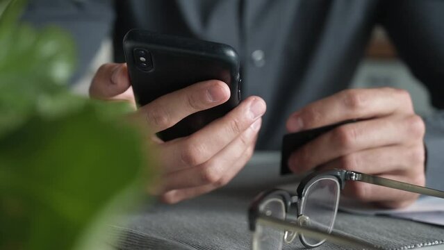 Close up cinematic shot of Man's hands dressed in black shirt holding a credit card and using smart phone for online shopping.

