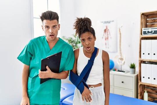 Physiotherapist working with patient wearing arm on sling at rehabilitation clinic in shock face, looking skeptical and sarcastic, surprised with open mouth