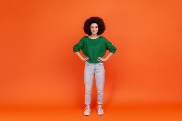 Fototapeta na wymiar Full length portrait of positive woman with Afro hairstyle wearing green casual style sweater standing with hands on hips and toothy smile. Indoor studio shot isolated on orange background.