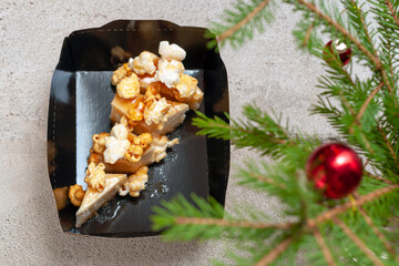 Obraz na płótnie Canvas cheesecake with salted caramel and popcorn in a black paper packaging box for delivery on a gray background with fir branches and Christmas balls.