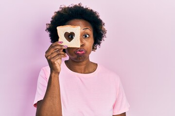 Young african american woman holding bread loaf with heart shape puffing cheeks with funny face. mouth inflated with air, catching air.