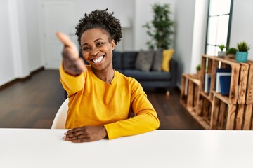 Young african american woman wearing casual clothes sitting on the table at home smiling friendly offering handshake as greeting and welcoming. successful business. - obrazy, fototapety, plakaty
