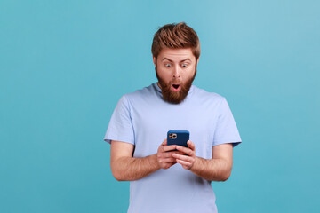 Portrait of astonished surprised young adult bearded man standing, using smartphone and watching video with amazed face and open mouth. Indoor studio shot isolated on blue background.