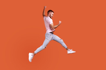 Fototapeta na wymiar Side view portrait of happy bearded man jumping in air showing yes i did it gesture, copy space for ad, wearing pink T-shirt and jeans. Indoor studio shot isolated on orange background.