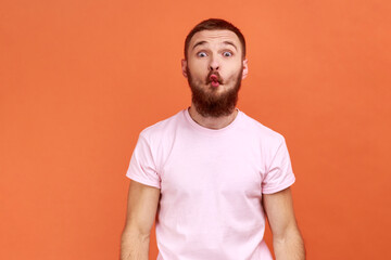 Portrait of funny childish bearded man making fish face or sending kiss, having fun, kidding, pretending to be stupid, wearing pink T-shirt. Indoor studio shot isolated on orange background.