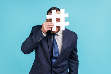 Man wearing official style suit hiding behind white hashtag symbol, covering face with hash sign,...
