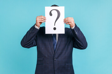 Portrait of anonymous male wearing dark official style suit covering his face with paper with question mark, finding right solution. Indoor studio shot isolated on blue background.