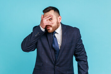 Bearded handsome man wearing official style suit looking through fingers, peeking with curious face, having suspicious, watching secrets. Indoor studio shot isolated on blue background.