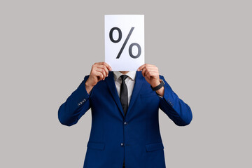 Portrait of anonymous unknown businessman hiding his face behind paper with percent sign inscription, wearing official style suit. Indoor studio shot isolated on gray background.