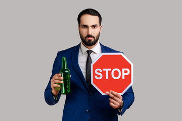 Portrait of serious strict bearded man showing alcoholic beverage beer bottle and stop sign,...