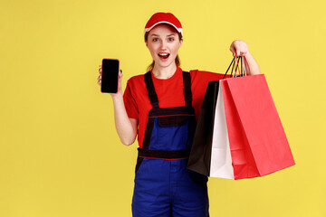Portrait of surprised courier woman standing with shopping bags in hands and showing smart phone with blank screen for ad, wearing overalls. Indoor studio shot isolated on yellow background.