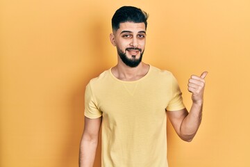 Handsome man with beard wearing casual yellow t shirt smiling with happy face looking and pointing to the side with thumb up.