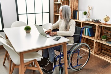 Middle age grey-haired woman uisng laptop sitting on wheelchair at home