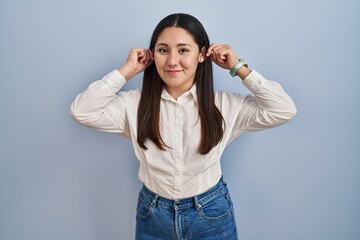 Young latin woman standing over blue background smiling pulling ears with fingers, funny gesture....