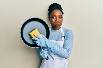 African american woman with braided hair wearing apron holding scourer washing pan puffing cheeks...