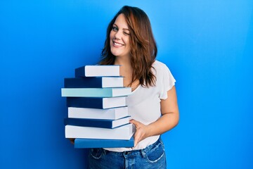 Young caucasian woman holding a pile of books winking looking at the camera with sexy expression, cheerful and happy face.