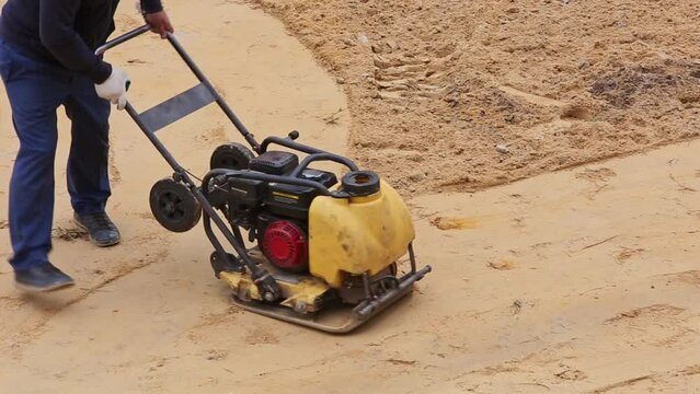 worker with vibration plate compactor ramming sand on construction site before paving