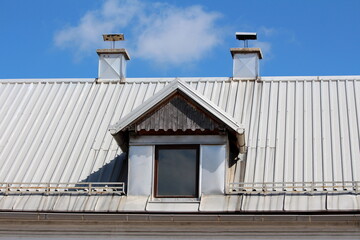 Old roof window with wooden frame and wooden dilapidated decorative boards on top covered with newly installed metal roof tiles surrounded with snow guards and two metal sheets covered chimneys on top