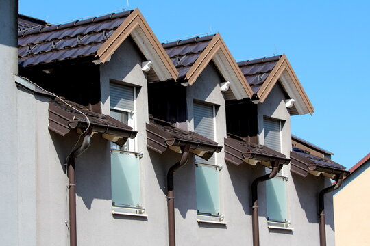 Row of three renovated old style roof windows covered with new dark brown roof tiles and metal lightning rod on top with opaque frosted glass balcony covers for added privacy on lover parts