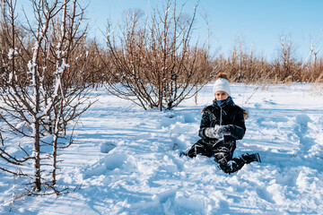 A little girl squints from the sun and sits in the snow in the winter in the forest