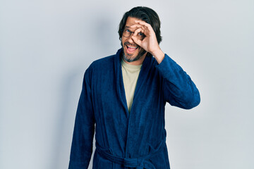 Middle age caucasian man wearing bathrobe and glasses doing ok gesture with hand smiling, eye looking through fingers with happy face.