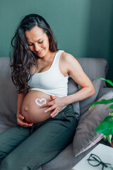 Calm pregnant woman with long dark hair in white shirt sitting on gray sofa with cream heart paint on tummy. Humidifier and table with plants. Moisturising, skin care, stretch marks prevention 