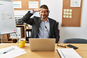 Handsome middle age man wearing call center agent headset at the office very happy and smiling looking far away with hand over head. searching concept.