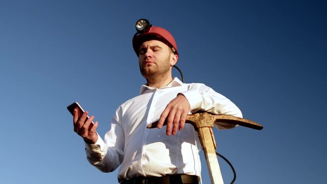 Businessman standing in helmet leaning on pickaxe and holding smartphone in hand, bottom view. Cryptocurrency miner outdoor with phone at hand and leans on pickaxe. Concept of cryptocurrency mining