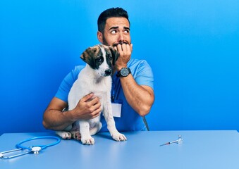Handsome hispanic veterinary man with beard checking dog health looking stressed and nervous with...