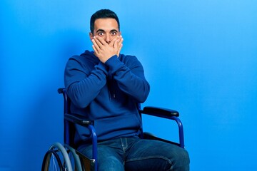 Handsome hispanic man with beard sitting on wheelchair shocked covering mouth with hands for mistake. secret concept.