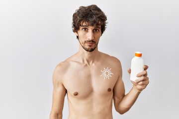 Fototapeta premium Young hispanic man standing shirtless holding sunscreen lotion relaxed with serious expression on face. simple and natural looking at the camera.