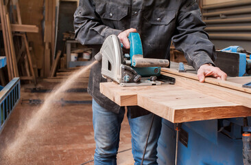 Male carpenter sawing a board with a circular saw in a carpentry workshop close-up