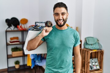 Young handsome man with beard holding shopping bags at retail shop winking looking at the camera with sexy expression, cheerful and happy face.