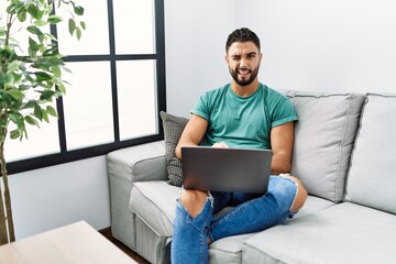 Young handsome man with beard using computer laptop sitting on the sofa at home winking looking at the camera with sexy expression, cheerful and happy face.