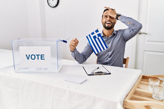 Young handsome man with beard at political campaign election holding greece flag stressed and frustrated with hand on head, surprised and angry face